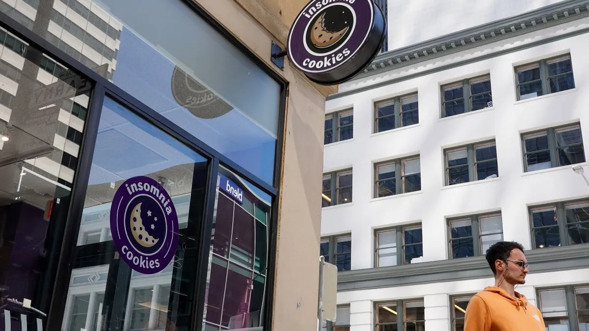 A man in an orange sweatshirt next to a corner of a building that has a circle sign with the words "insomnia cookies."