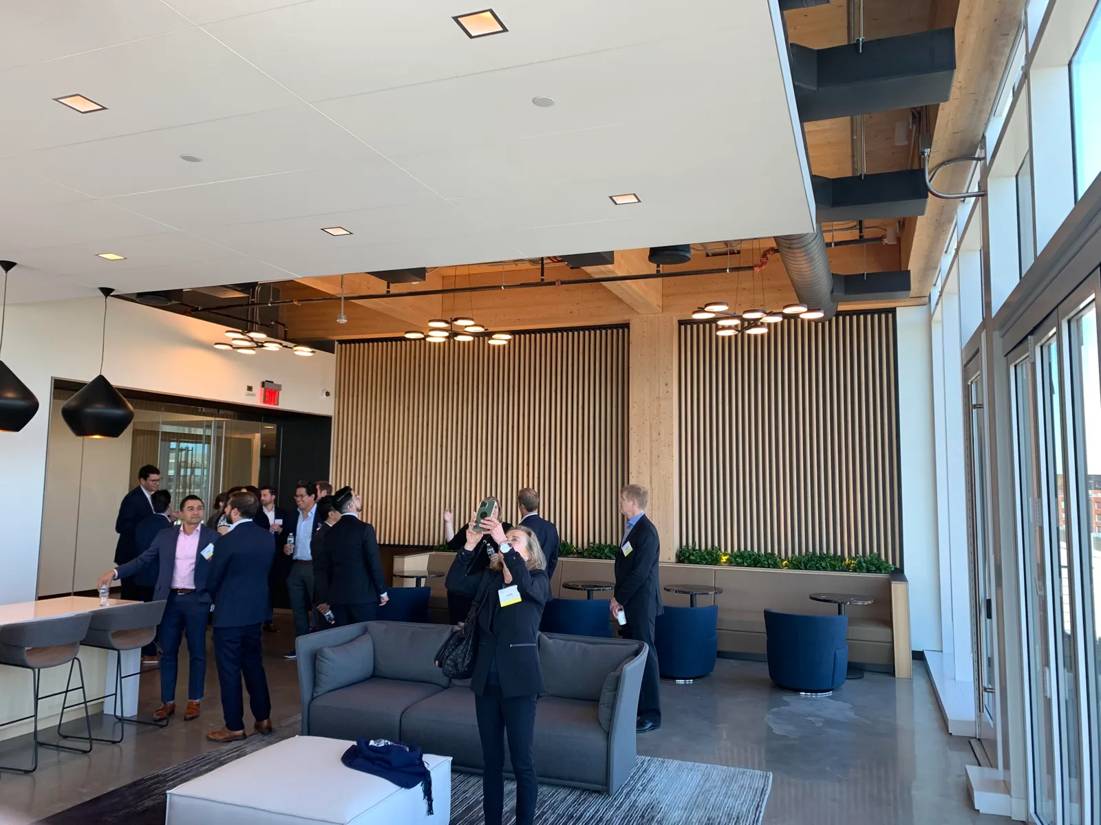 A room with wooden columns and wood paneling is in the background and foreground. People in business attire mill about, chatting and taking pictures.