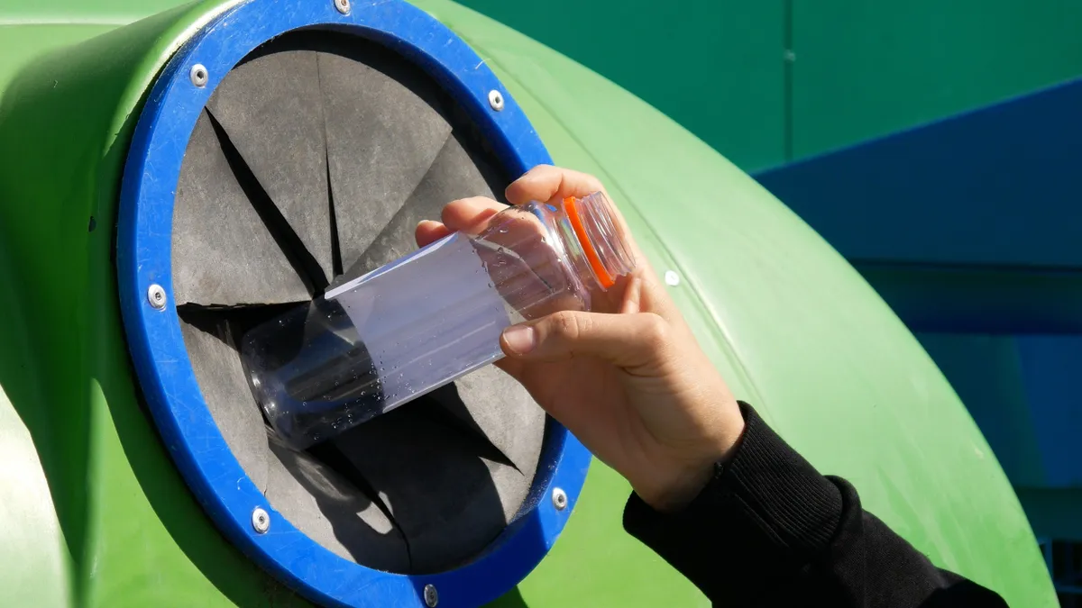 A close up of a hand throwing away a plastic bottle into a recycling container.