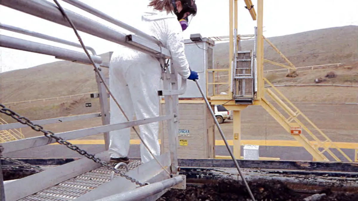 A person wearing a hazmat suit stands on a platform holding a long pole reaching into a dumpster. A mound rises behind them.