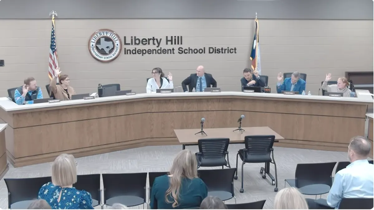 Seven people sit behind a dias and six of them are raising their hands. Behind them is a sign reading Liberty Hill Independent School District.