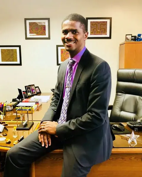 A person is leaning/sitting on a desk in an office. The person is looking at the camera.