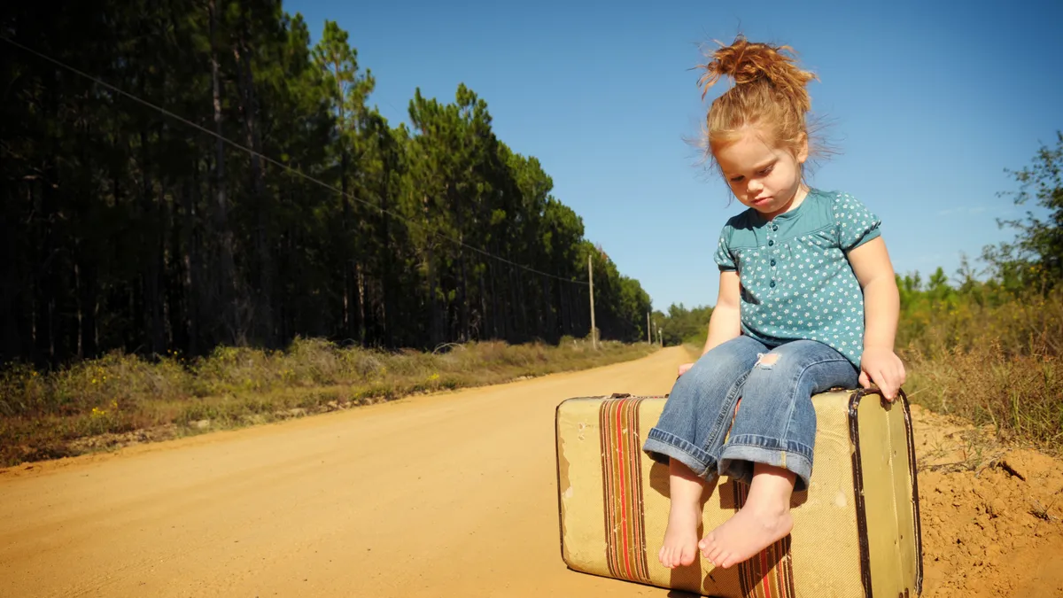 A child sits on a suitcase in the middle of a dirt road