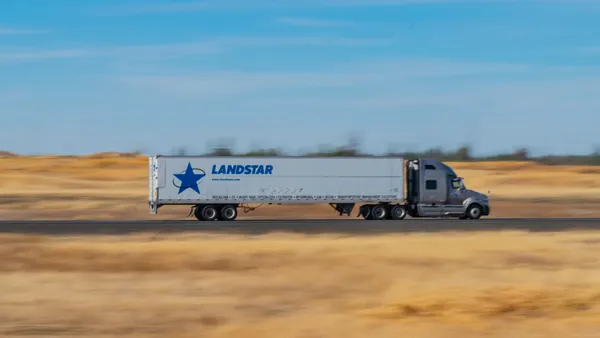 A Landstar System semi-truck driving along a rural section of an interstate with blue sky and wispy clouds in the background.