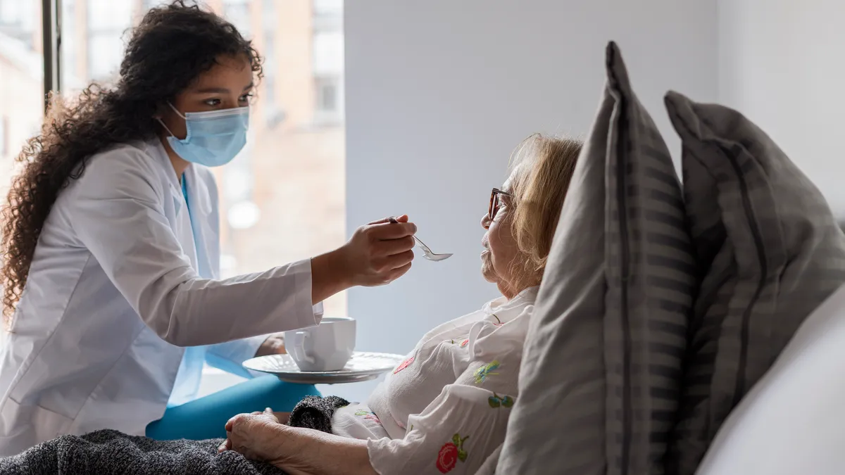 A home health aide sits on a bed and uses a spoon to feed an older individual lying in the bed.