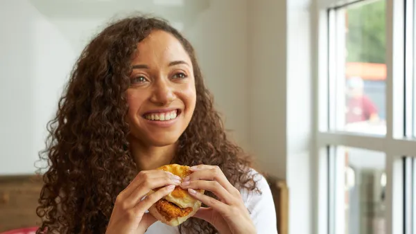 A photo of a person sitting next to a window inside a building, holding a breaded chicken sandwich.