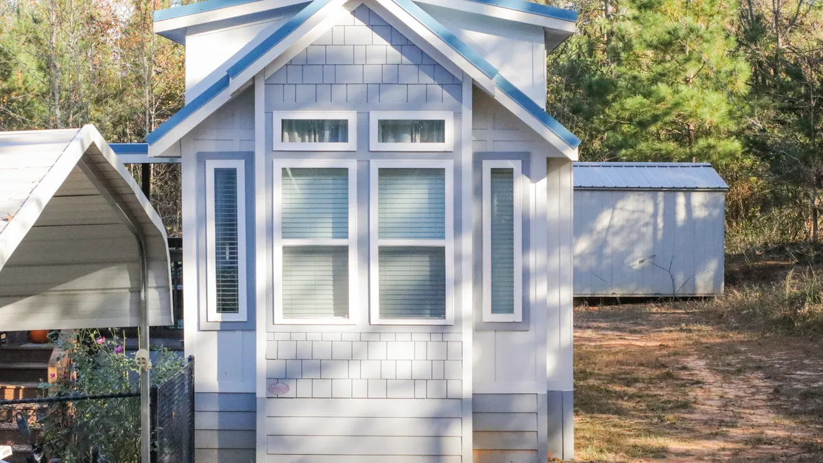 Front end of a blue tiny house with under skirting and storage unit in the back. Shaded windows in the front of tiny home. Fence.