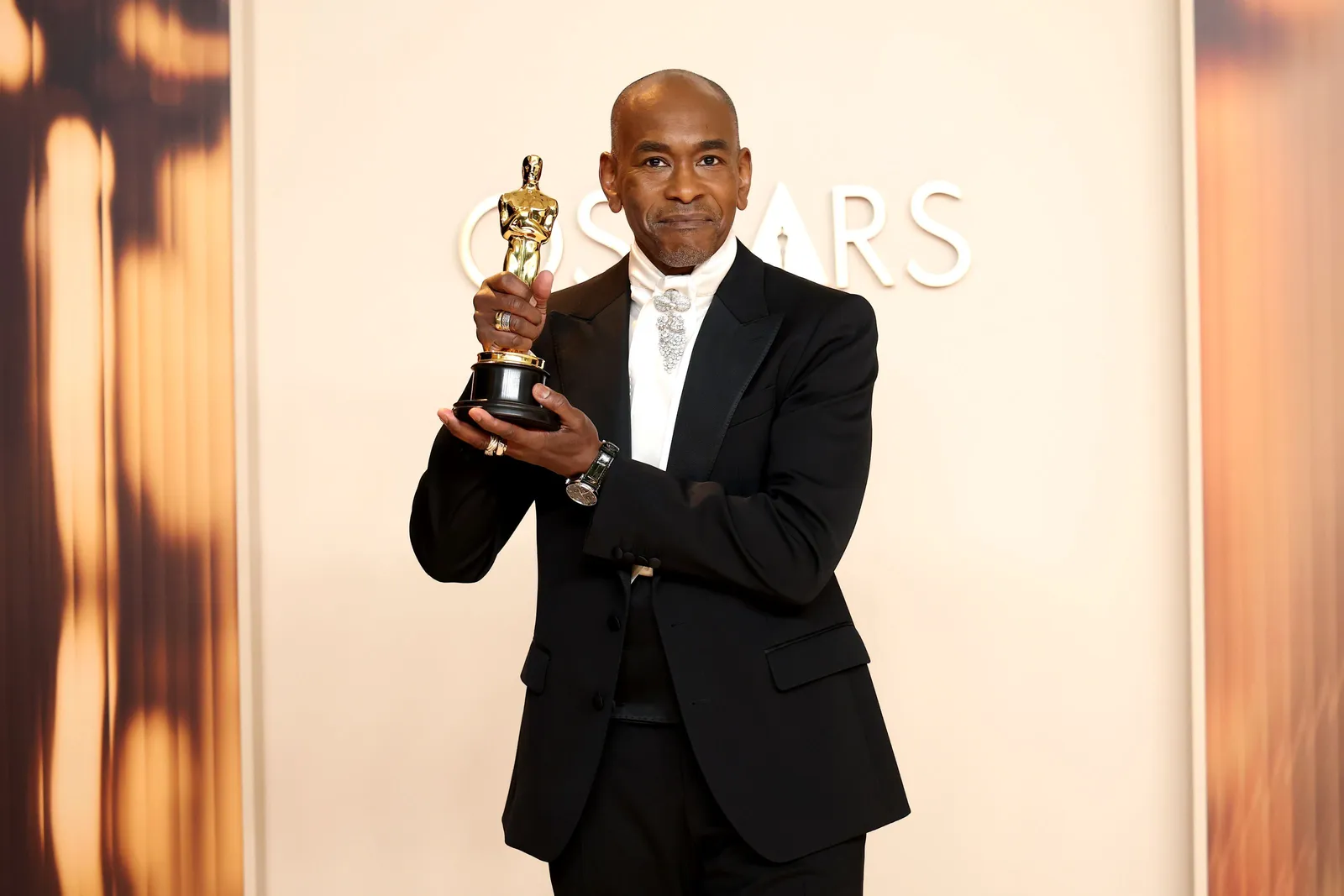 A man in a classic tuxedo and cumberbund accentuated with a white ascot and a diamond brooch stands on the red carpet holding an Oscar.