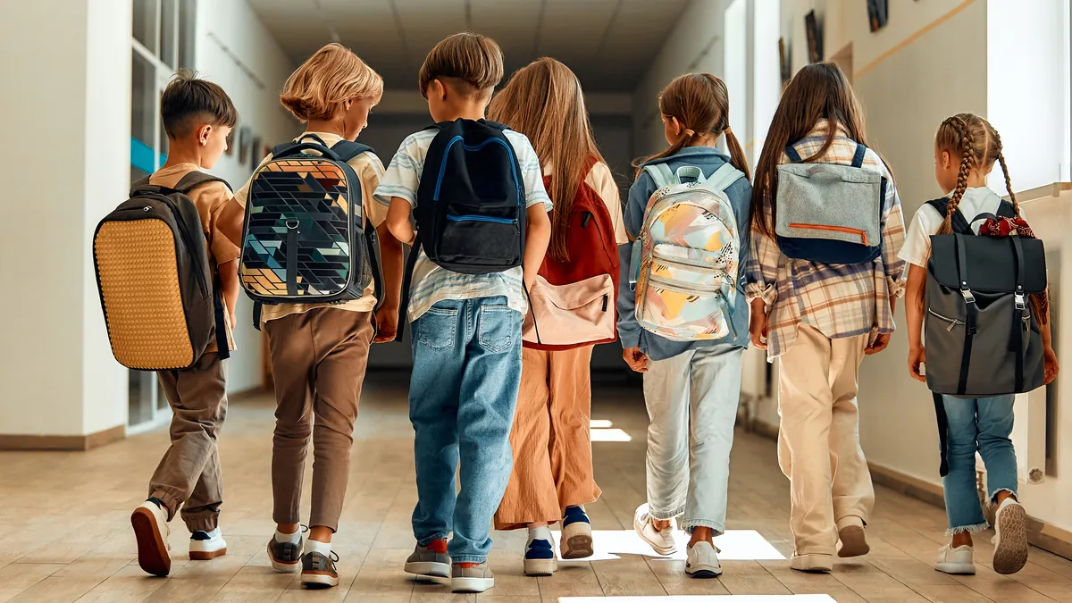 Six students carrying backpacks are walking down a hallway in a school. Their backs are to the camera and light is coming in through a window.