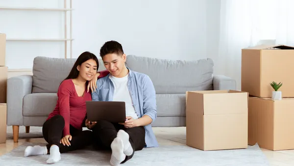 2 People sitting in a room in front of a couch surrounded by moving boxes and looking at a tablet.