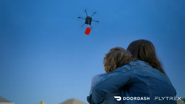 An image of an adult and child looking up at the sky where there is a drone carrying a red bag.