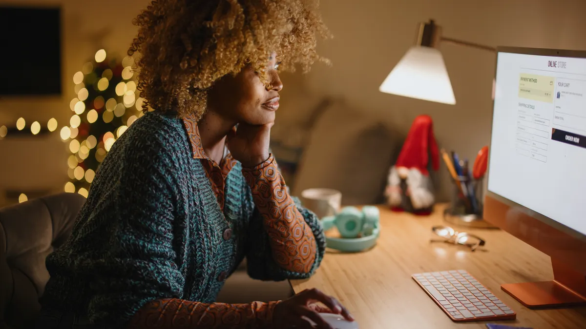 A person sits in front of a computer online shopping while a Christmas lights are in the background.