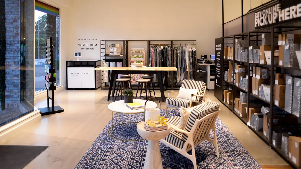 Wide angle photo showing the interior of a storefront with seating in the foreground and clothes on hangers in the background, plus clothing shopping bags on the right for online order pickups.
