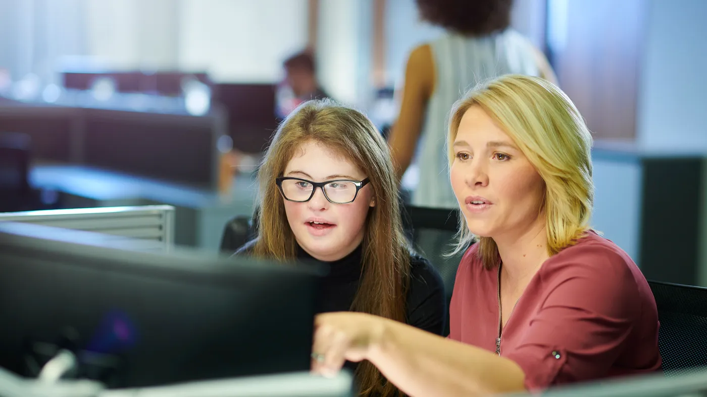 A high school student sits at a desk with an adult. They are looking at a computer screen