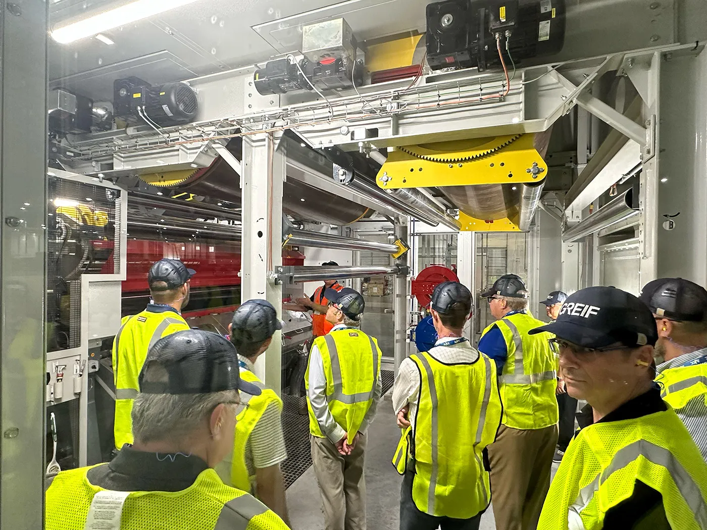 A group of people wearing yellow vests and protective glasses listen to a speaker in an industrial facility.