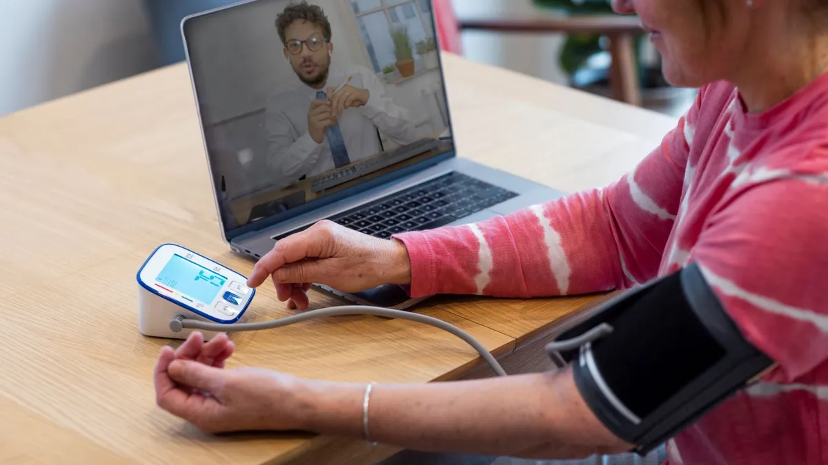 Woman using a laptop to contact a medical professional about her health. She is siting at a table in a kitchen and is taking her blood pressure with a blood pressure gauge.