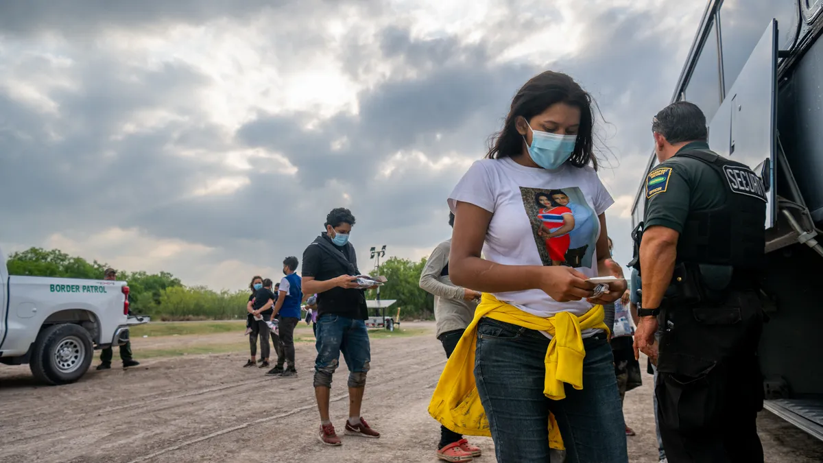 Masked people in La Joya, Texas line up under a cloudy sky, preparing to board a U.S. Border Patrol bus