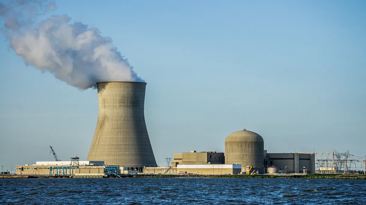 Steam billows from a massive cement smokestack at a nuclear power plant at the edge of the water.