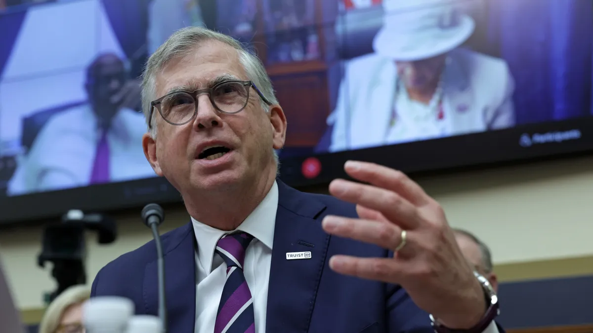 Bill Rogers, wearing a blue suit jacket, speaks during a Congressional hearing, with a screen behind him