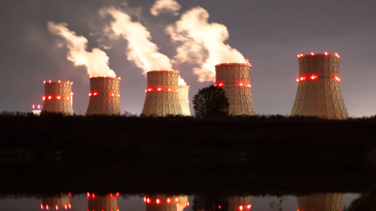 Multiple cooling towers illuminated with red lights emit large plumes of steam into the night sky. The towers are reflected in a still body of water in the foreground, with a silhouetted tree and landscape between the towers and the water.