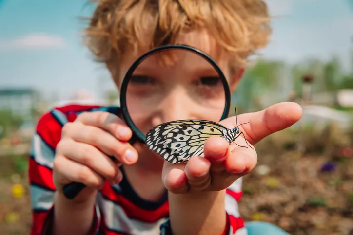 A student is holding a butterfly outside. The student has a magnifying glass to their face.