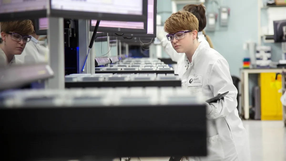 A person wearing goggles and holding a clipboard looks at a tray in a laboratory.