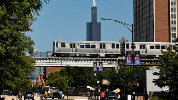 A silver elevated train over a city street with a tall building in the background.