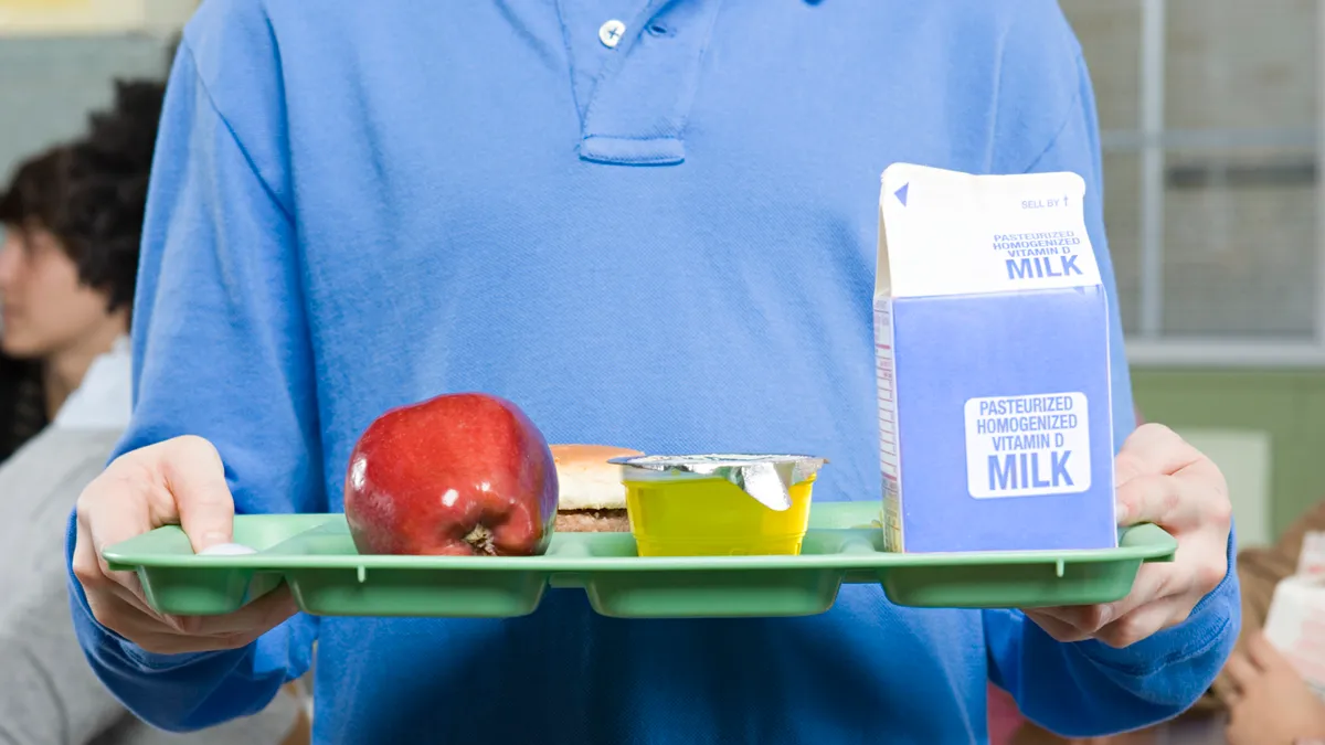 A student holds a tray of food and a milk carton as students sit behind him in a school cafeteria.