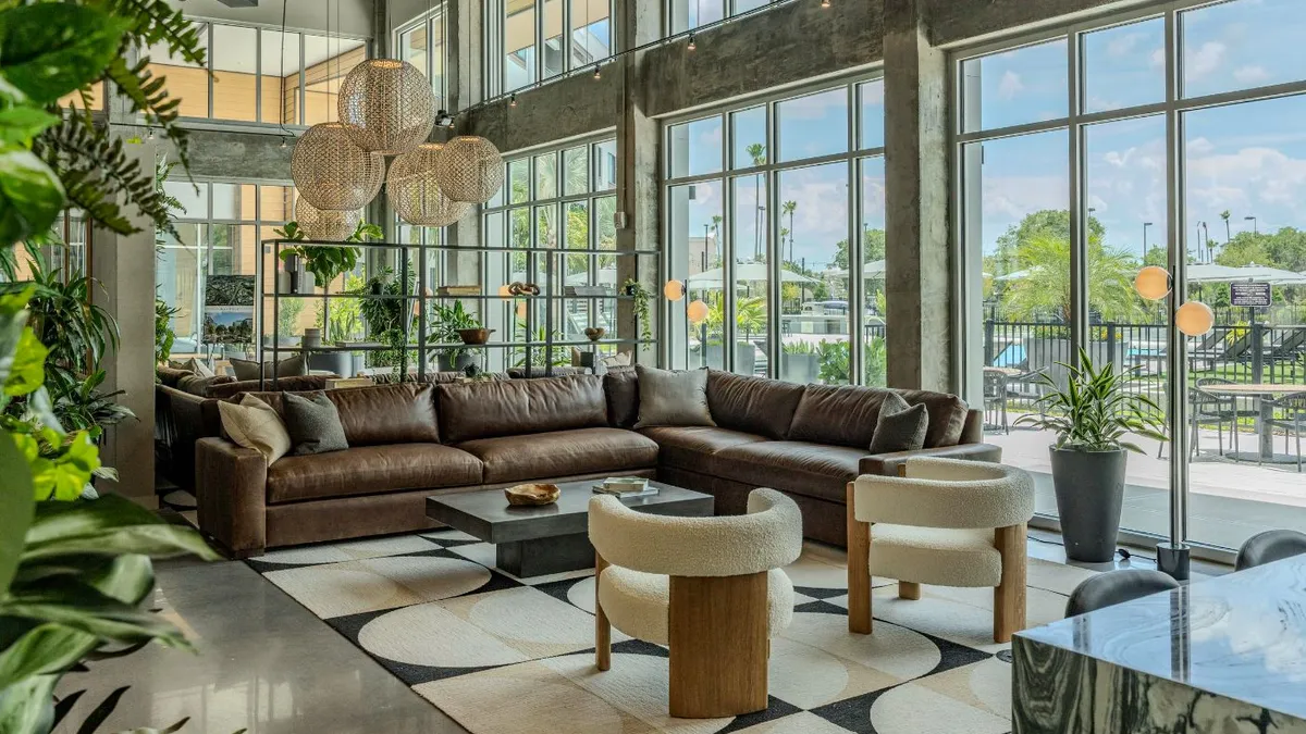 A lounge area with tall glass windows. A pool and tall trees are visible outside.