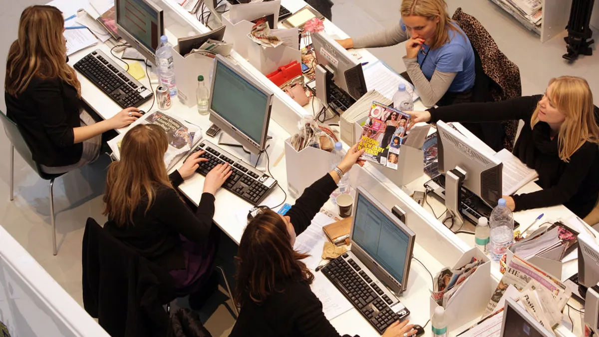 Women sit at computers together at a long table working on projects.