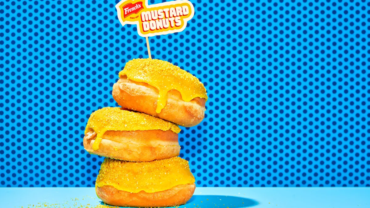 Three of French's yellow mustard-flavored donuts are stacked vertically on a stick, promoting the release of the themed donuts.