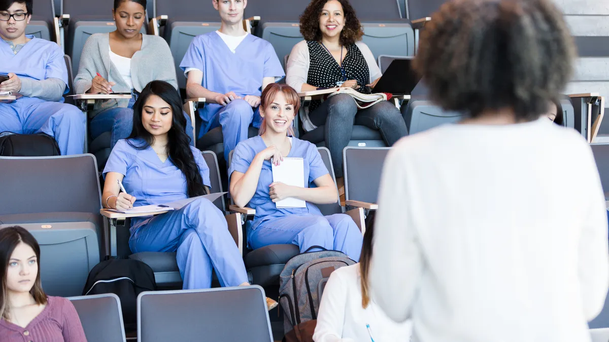 People in scrubs and street clothes sit in an auditorium in front of a lecturer.