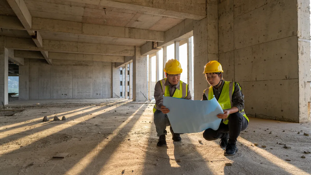 Two construction engineers with yellow hard hats looking at an unrolled white paper in an empty buidling with dirt floor, and sun rays coming through the building's openings.