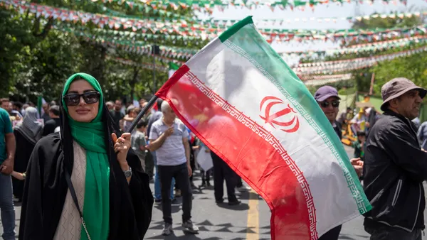 A woman wearing a hijab and sunglasses waves an Iranian flag