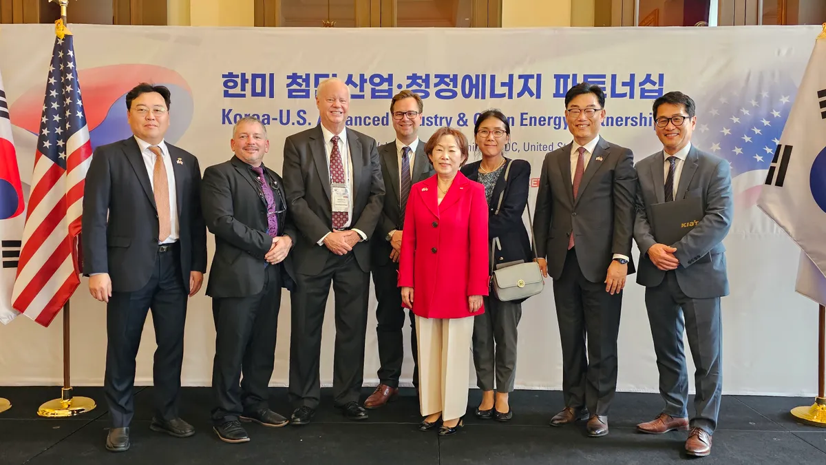 A group of people standing in front of a large white banner with blue Korea text on top and english text underneath. South Korea and U.S. flags are on the left and right.