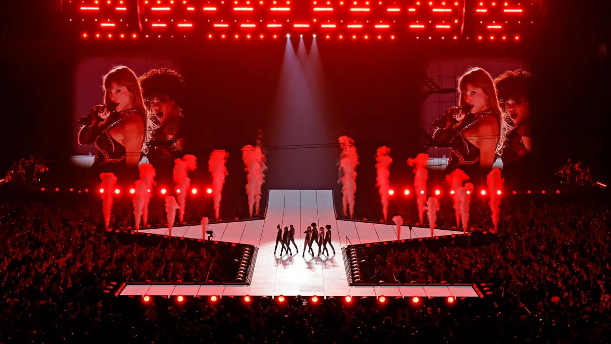 Taylor Swift performing live onstage flanked by dancers in front of a stadium crowd. In the background are bursts of smoke and large screens featuring images of Swift.