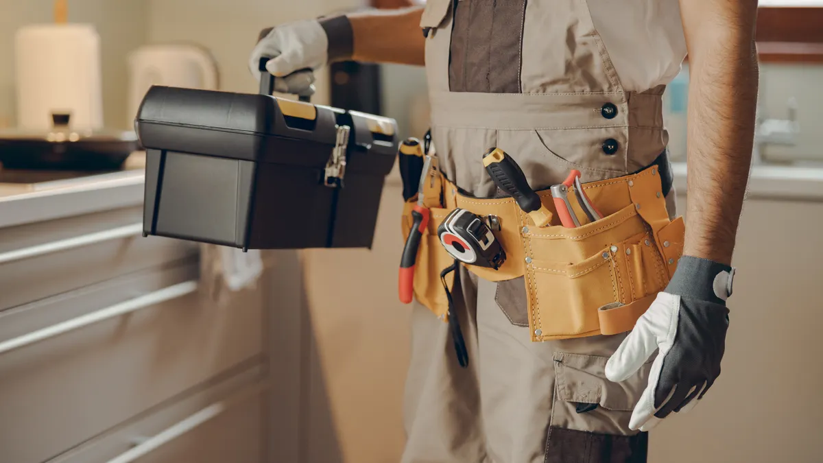 Close up of repairman in uniform standing on home kitchen and holding his tool bag