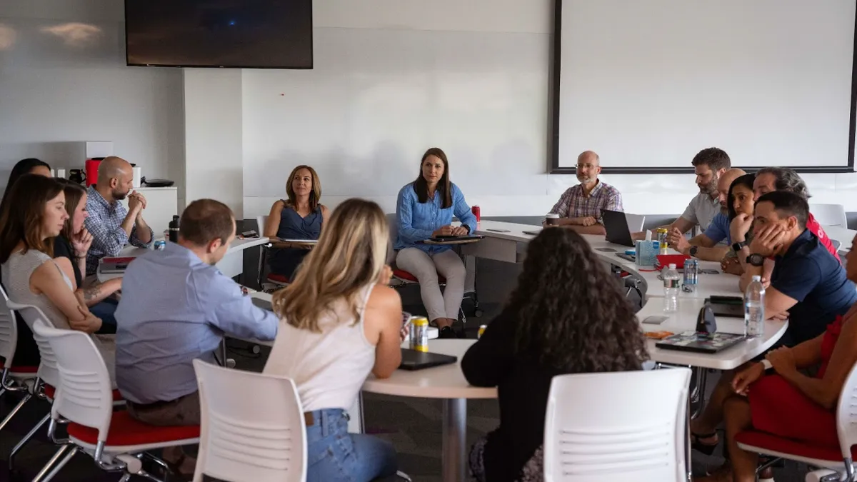 A group of people in a large office meeting room sitting around a circle of tables.