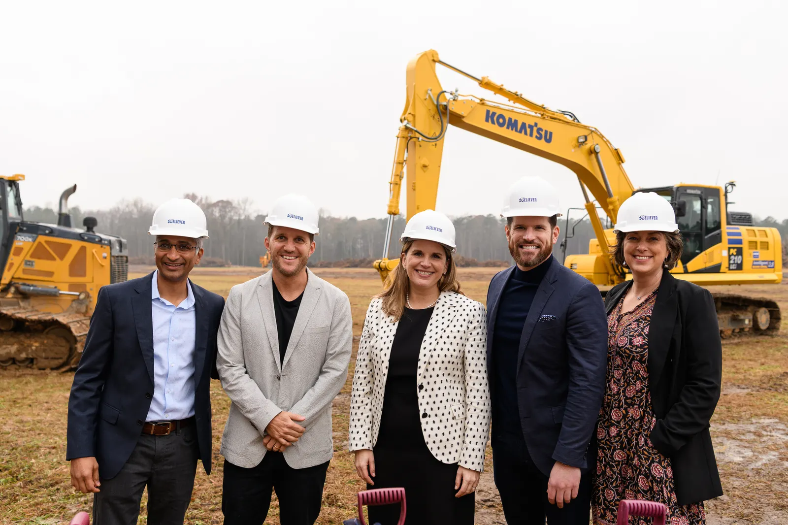 A group of well dressed people in white hard hats stand on a cleared lot with construction equipment in the background