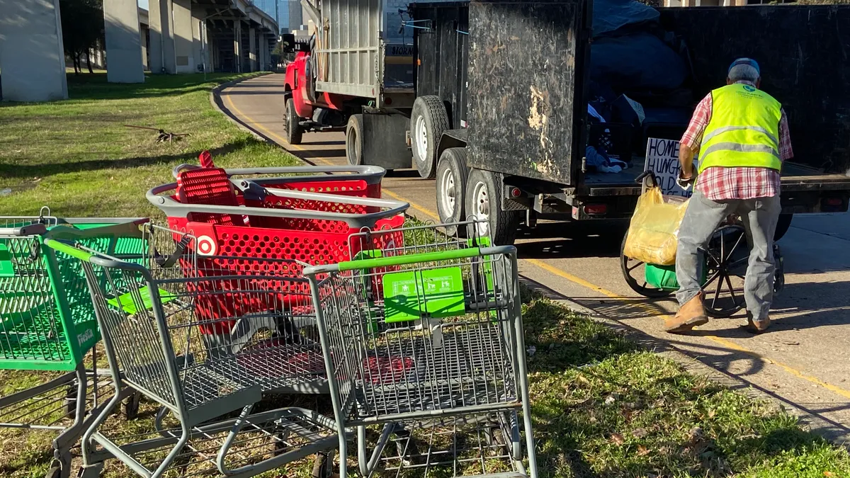 A person in a yellow vest loads items into the pack of a truck parked below a raised freeway. A group of shopping carts sit on the grass nearby.