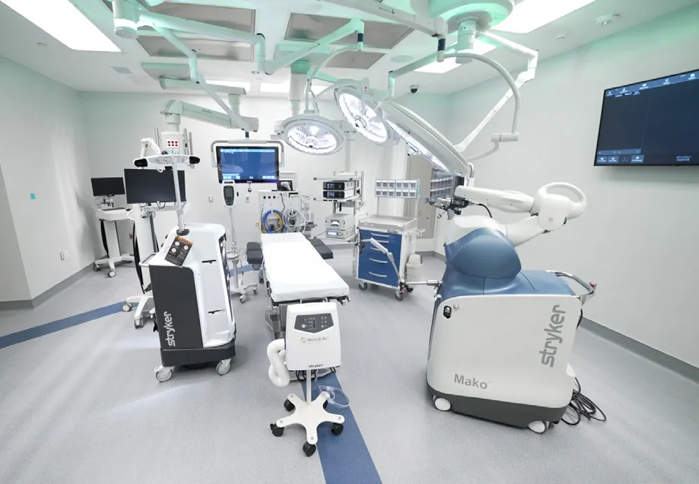 An operating room has a table surrounded by screens and several overhead lights, and a surgical robot that says "Mako"