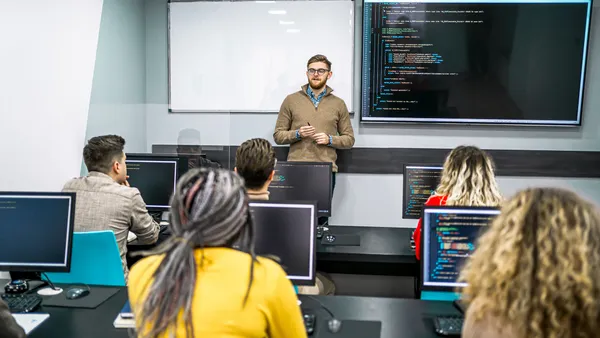 An instructor stands at the front of a classroom talking to several students sitting at desks.