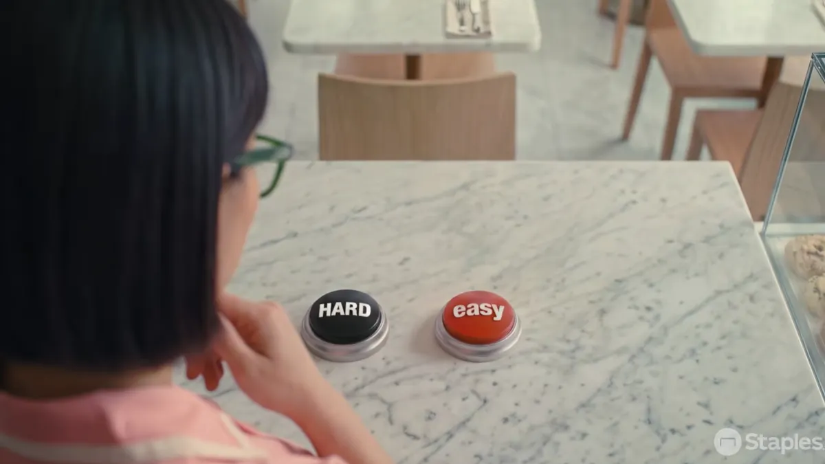 A person faces a black Hard Button and red Easy Button on a desk.