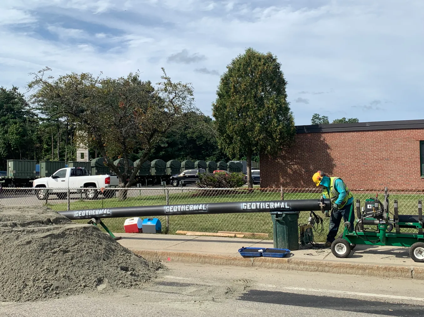 A worker in a hard hat and yellow vest lays a large pipe in the ground. On the side of the pipe, the word "geothermal" is written three times.