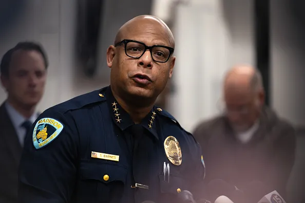 An adult in a police uniform is speaking in front of a row of microphones. Two other  people are seen in the background.