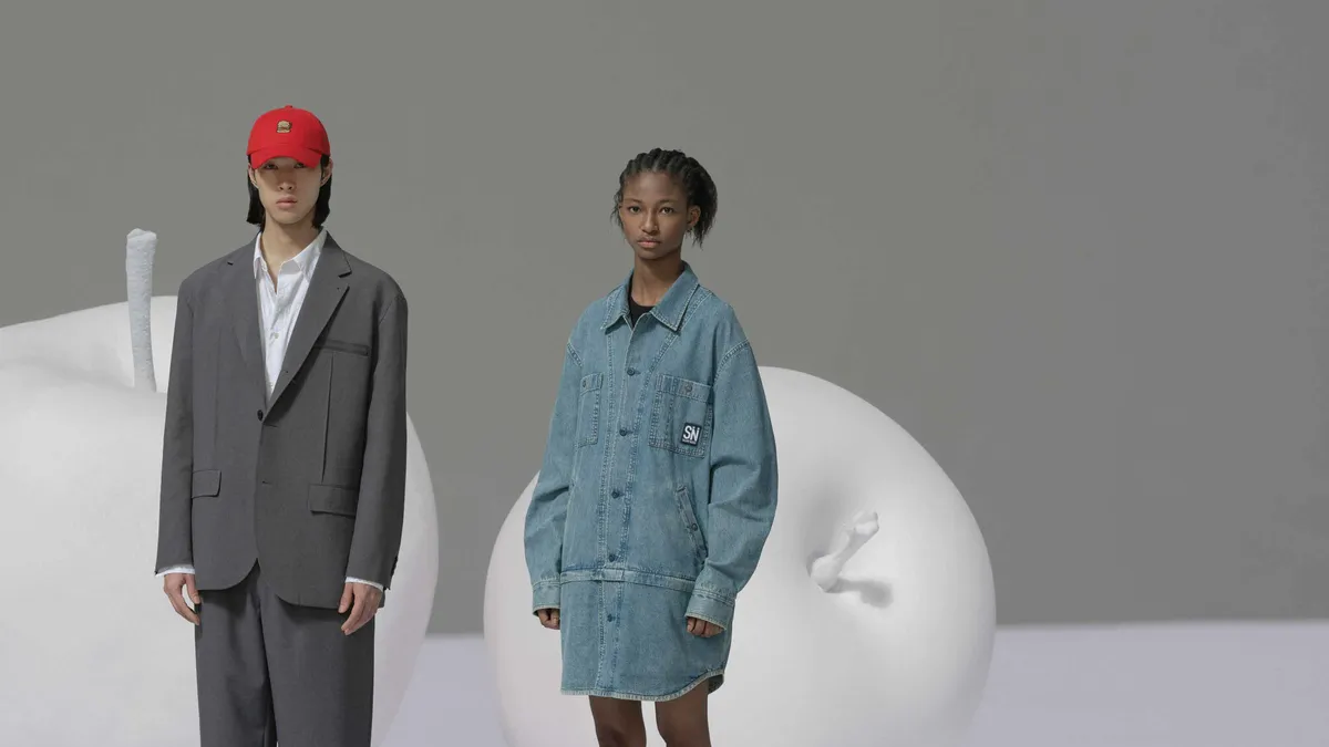 Two people stand in front of large white sculptures of apples. One wears a gray suit and the other wears a denim dress.