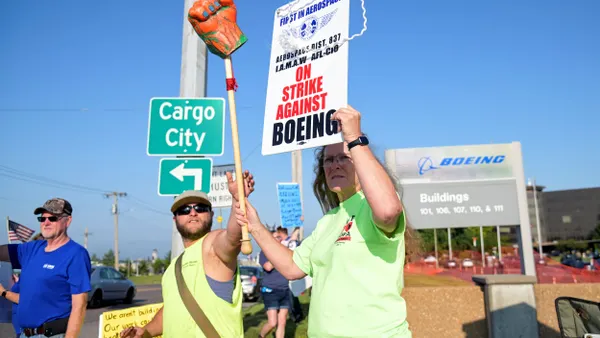 Three people holding up strike signs in front of a Boeing sign.
