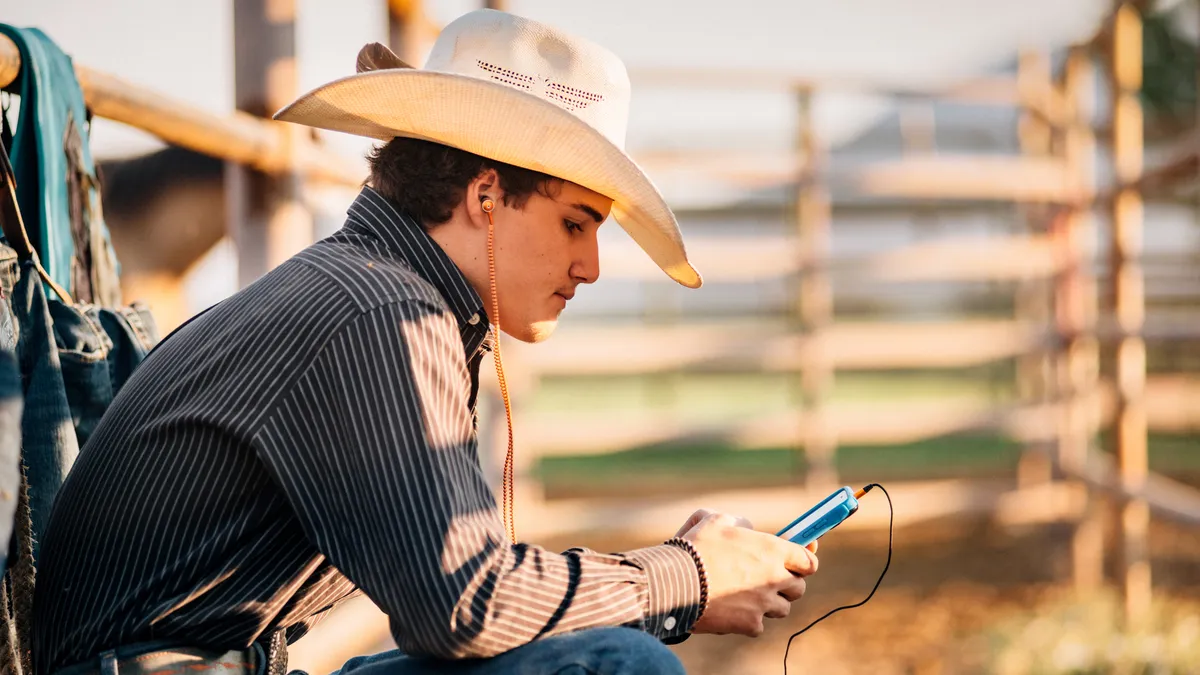 A rancher in a cowboy hat wears headphones and looks at his phone.