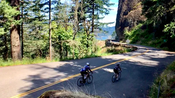 Two people on bicycles along a two-lane road surrounded by trees.