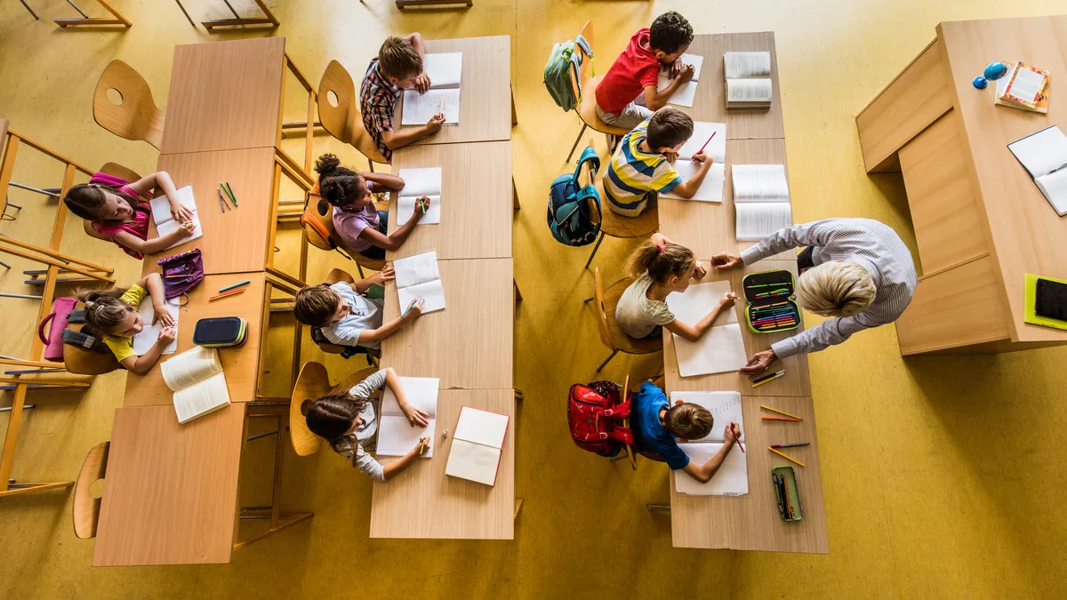 A view from above of an elementary classroom with rows of desks and students sitting at the desks with notebooks on the desks. An adult is standing in front of the desks helping a student.